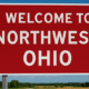 A red highway sign reading "Welcome to Northwest Ohio" stands against a bright blue sky with scattered clouds, symbolizing entry into the region.