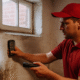 Maintenance technician in a red Buckeye Northwest Realty uniform inspecting a damp basement wall with a moisture meter and flashlight in an older Toledo home.