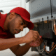 Maintenance technician in a red Buckeye Northwest Realty uniform lying on the floor to repair the back of a stainless steel refrigerator in a modern rental kitchen.